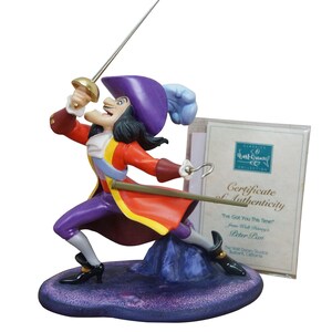 May include: A Captain Hook figurine in a dynamic fencing pose, wearing a red coat, purple hat with a blue feather, and black boots. The character holds a sword and has a metal hook hand. A Certificate of Authenticity is included in a plastic sleeve.