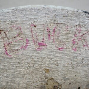 May include: Close-up of a weathered, white-painted wooden surface. The word "BLACK" is hand-painted in pink, with visible texture and imperfections. The wood grain is prominent, and the paint is chipped.