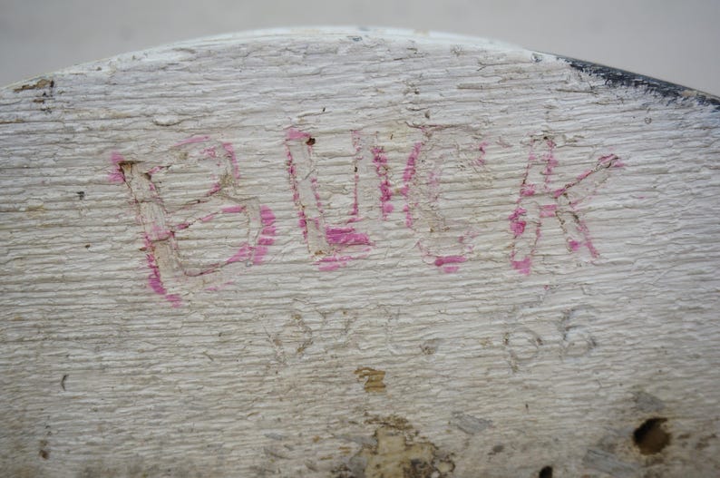 May include: Close-up of a weathered, white-painted wooden surface. The word "BLACK" is hand-painted in pink, with visible texture and imperfections. The wood grain is prominent, and the paint is chipped.