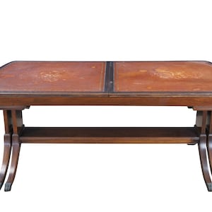 May include: A rectangular wooden coffee table with a brown leather top. The table has a lower shelf and curved legs. The leather top is divided into two sections and has a decorative design. The table is a classic piece of furniture.