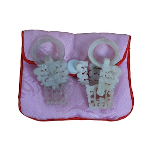 May include: Two carved jade pendants with ring handles and intricate designs, displayed on a pink pouch with red trim. The pendants are a pale green color, and the pouch has a soft, textured appearance.