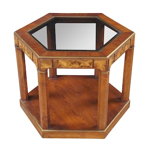 May include: An octagonal wooden side table with a glass top and a lower shelf. The table is made of a light brown wood with a darker wood grain pattern. The edges are trimmed with a gold-colored metal.
