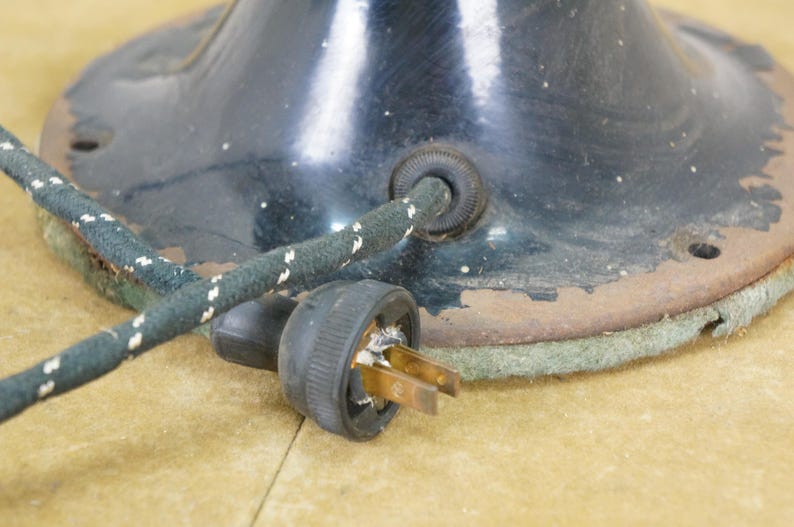 Puede incluir: Primer plano de una base de l&aacute;mpara antigua de metal negro con cable de alimentaci&oacute;n y enchufe. La base tiene un dise&ntilde;o circular con un borde oxidado. El cable es verde oscuro con motas blancas. El enchufe tiene dos clavijas de lat&oacute;n.