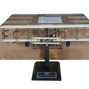 May include: A detailed model of a biplane, featuring a cream-colored wing structure with brown support struts. The model is mounted on a black stand with a nameplate. The biplane is positioned in front of a cardboard box.