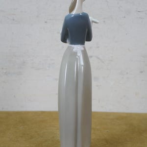 May include: A porcelain figurine of a woman, viewed from the back. She wears a gray dress, a blue top, and a white apron. Her hair is pulled back. The figurine stands on a light green base.