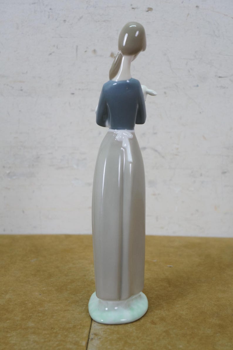 May include: A porcelain figurine of a woman, viewed from the back. She wears a gray dress, a blue top, and a white apron. Her hair is pulled back. The figurine stands on a light green base.