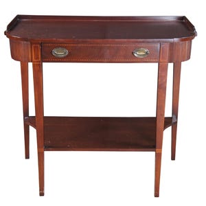May include: A mahogany-colored antique side table with a drawer and a lower shelf. The table has a rectangular top with rounded edges and a small drawer with two brass pulls. The legs are slender and tapered, and the table has a decorative inlay.