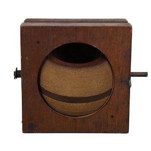 May include: A wooden box with a round, brown and tan object inside. The object is surrounded by a wooden frame and has a small metal knob on the side.