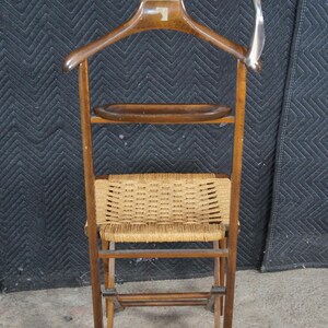 May include: A vintage wooden valet stand with a built-in hanger, a small shelf, and a woven seat. The stand is made of dark wood and features a silver-colored metal hook. The woven seat is made of a light brown material.