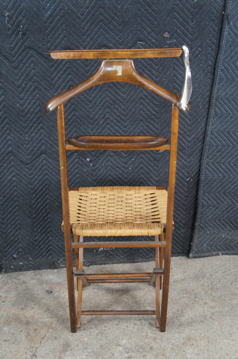 May include: A vintage wooden valet stand with a built-in hanger, a small shelf, and a woven seat. The stand is made of dark wood and features a silver-colored metal hook. The woven seat is made of a light brown material.