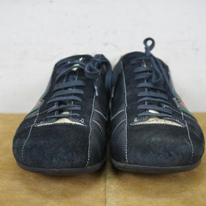 May include: A pair of navy blue suede sneakers with white stitching and matching laces. The shoes have a low profile design with a logo on the tongue and a green and red stripe detail on the side.