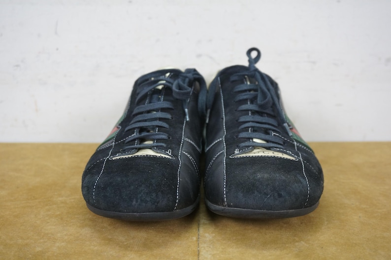 May include: A pair of navy blue suede sneakers with white stitching and matching laces. The shoes have a low profile design with a logo on the tongue and a green and red stripe detail on the side.