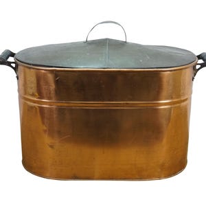 May include: An oval copper tub with a dark metal lid featuring a central handle. The tub has two side handles with dark grips. The copper body has a smooth, reflective surface with horizontal ridges near the top rim and a flat base.