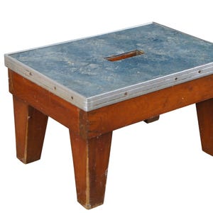 May include: A wooden stool with a blue metal top and a rectangular cutout in the center. The stool has four legs and a silver metal trim around the top.