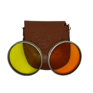 May include: Two round camera filters in a brown leather case. The filters are in silver frames and have a yellow and orange tint.