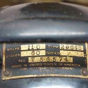 Puede incluir: Primer plano de una placa de motor el&eacute;ctrico antiguo. La placa dorada tiene texto negro que indica 110 voltios, 60 ciclos y el tipo 29648. El texto tambi&eacute;n dice "Made in United States of America for Alternating Current."