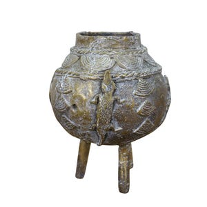 May include: A bronze-colored, three-legged decorative pot with intricate patterns and a lizard-like figure. The pot has a rounded body and a textured surface, suggesting an aged or antique appearance. The design includes circular and linear patterns.