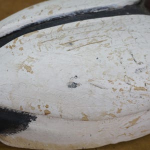 May include: Close-up of a weathered, painted duck decoy. The decoy features a white body with black and brown accents. The paint is distressed, showing signs of age and use. The duck's head is a dark maroon color.