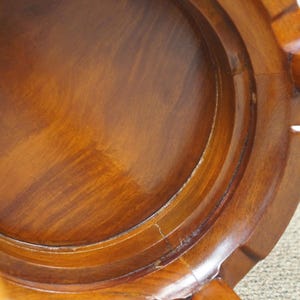 May include: Close-up of a wooden stool, showcasing the underside with a circular seat and curved, layered design. The wood has a warm, reddish-brown hue with visible grain and a glossy finish. The legs are visible.