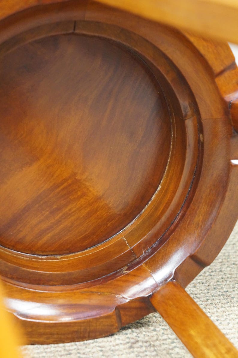 May include: Close-up of a wooden stool, showcasing the underside with a circular seat and curved, layered design. The wood has a warm, reddish-brown hue with visible grain and a glossy finish. The legs are visible.