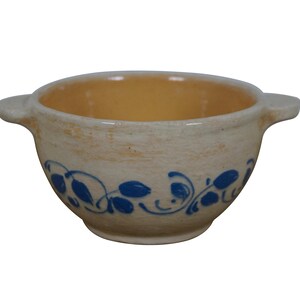 May include: A small, cream-colored ceramic bowl with blue floral designs. The bowl has two handles and a slightly rounded bottom.