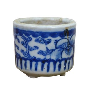 May include: A small, cylindrical ceramic container with a blue and white floral design. The container has a rounded top and a base with three small feet. The blue design includes floral and scroll patterns. The container is approximately 2.5 inches tall.