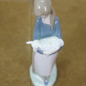 May include: A ceramic figurine of a woman in a blue dress holding a white lamb. The figurine has a light brown head and a long, light brown ponytail. The base is light green.