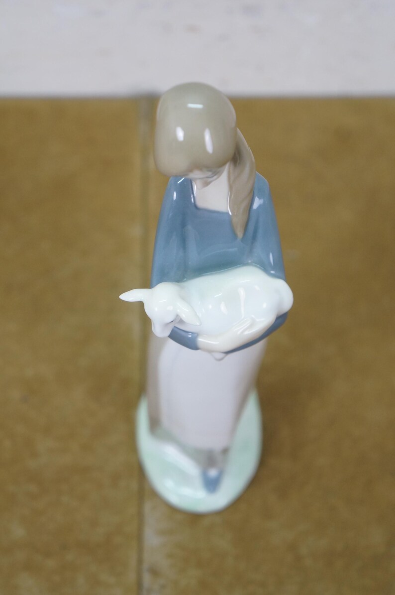 May include: A ceramic figurine of a woman in a blue dress holding a white lamb. The figurine has a light brown head and a long, light brown ponytail. The base is light green.