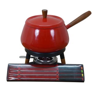 May include: A red fondue pot with a wooden handle and lid, set on a black stand with wooden legs. A box of fondue forks is in front of the pot. The pot is designed for cooking and serving.