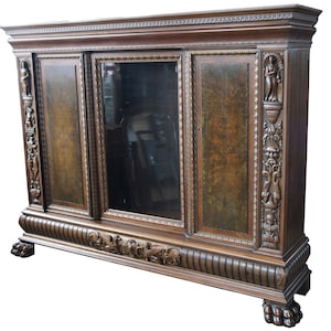May include: A large, dark brown wooden cabinet with intricate carvings and a glass door in the center. The cabinet has a decorative top and sits on four carved feet.