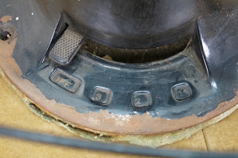Puede incluir: Primer plano del panel de control negro de un ventilador vintage. El panel presenta un interruptor deslizante y ajustes de velocidad numerados: Apagado, 3, 2 y 1. La carcasa met&aacute;lica del ventilador muestra signos de edad con &oacute;xido en los bordes.