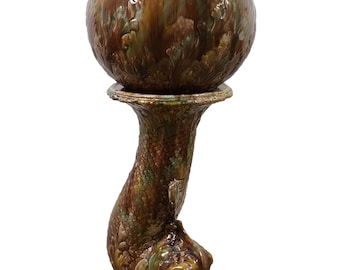 May include: A ceramic planter with a textured, ruffled rim, in shades of brown and green. The planter sits atop a pedestal shaped like a stylized dolphin, with a detailed, textured finish. The overall design is ornate and decorative.