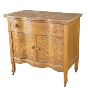 May include: A light brown wooden cabinet featuring a serpentine front, one wide top drawer with two round knobs, and two lower cabinet doors with matching round knobs. The piece stands on four legs equipped with small metal casters for mobility.