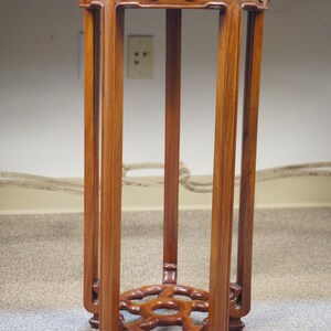 May include: A tall, wooden plant stand with a warm brown finish. The stand features a square top and base, connected by four vertical supports. Intricate carved details adorn the top and base, adding a touch of elegance. The stand is approximately 30 inches tall.