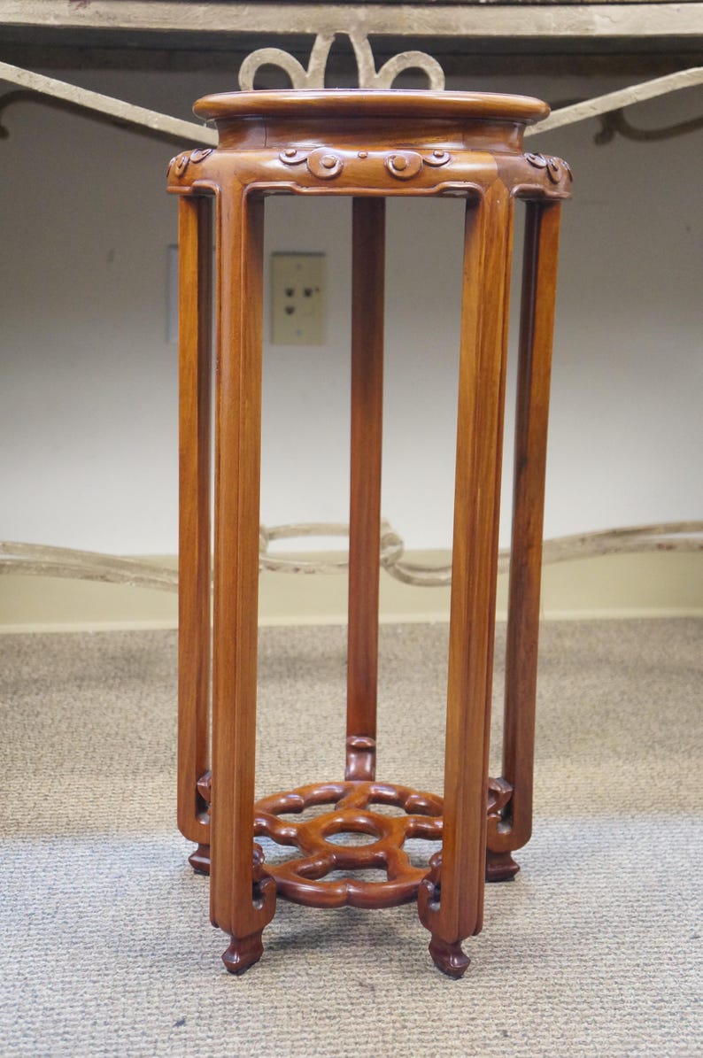 May include: A tall, wooden plant stand with a warm brown finish. The stand features a square top and base, connected by four vertical supports. Intricate carved details adorn the top and base, adding a touch of elegance. The stand is approximately 30 inches tall.