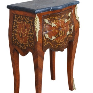 May include: A small, antique wooden nightstand with a dark marble top. The nightstand features two drawers with brass handles and decorative brass accents. The wood has a patterned design with inlaid details.