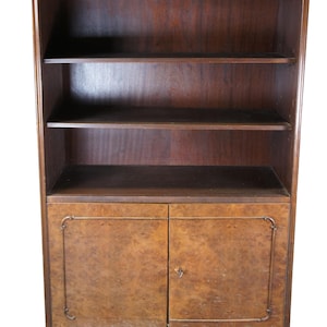 May include: A dark wood bookcase with three shelves and two lower cabinet doors. The cabinet doors have a lighter wood grain pattern and a decorative metal trim. The top of the bookcase has a carved design.