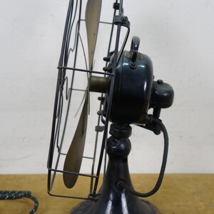 Puede incluir: Un ventilador el&eacute;ctrico negro antiguo con una jaula y tres aspas. El ventilador tiene una base redonda y un asa en la parte superior. El cable de alimentaci&oacute;n es blanco y negro. El ventilador es probablemente de principios del siglo XX.