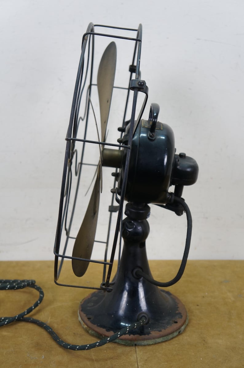 Puede incluir: Un ventilador el&eacute;ctrico negro antiguo con una jaula y tres aspas. El ventilador tiene una base redonda y un asa en la parte superior. El cable de alimentaci&oacute;n es blanco y negro. El ventilador es probablemente de principios del siglo XX.