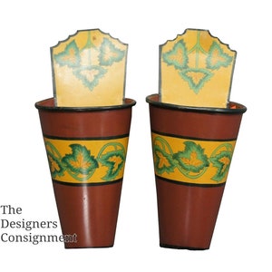 May include: Two brown metal wall planters with a yellow band featuring green leaf designs. Each planter has a yellow top with a green leaf design. The planters are against a white background.