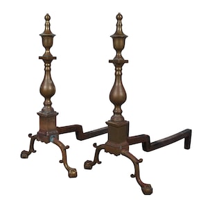 May include: A pair of antique brass fireplace andirons with ornate details. The andirons have a curved base and a tall, slender design with a decorative finial.