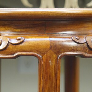 May include: Close-up of a wooden plant stand with a rich, warm brown finish. The stand features carved scrollwork along the edges and sturdy legs. The stand's design is classic and elegant, suitable for displaying plants or decorative items.