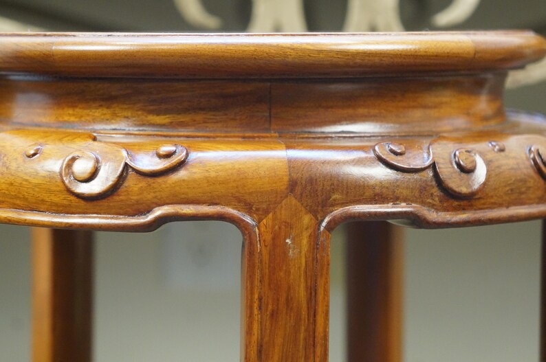 May include: Close-up of a wooden plant stand with a rich, warm brown finish. The stand features carved scrollwork along the edges and sturdy legs. The stand's design is classic and elegant, suitable for displaying plants or decorative items.