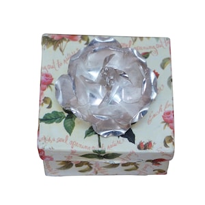 May include: A square gift box with a floral design and a large silver rose on top. The box has a cream background with pink roses and green leaves. The silver rose is the focal point.