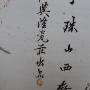 Op de afbeelding: Close-up van een witte keramische vaas met zwarte kalligrafie in Chinese karakters. De karakters lezen "春 天 曉 霞 飛 舟", wat vertaalt naar "Lente ochtend, wolken vliegen, boot". Er is een rode stempel op de vaas.