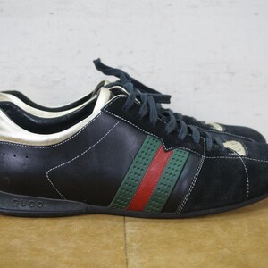May include: Black and gold sneakers with green and red stripes. The shoes have black laces and the brand name "Gucci" is visible on the sole. The shoes are made of leather and suede.