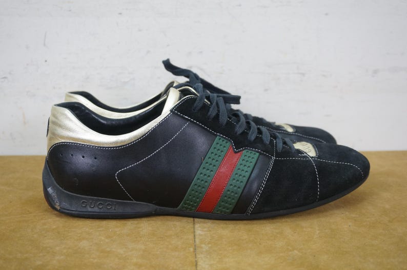 May include: Black and gold sneakers with green and red stripes. The shoes have black laces and the brand name "Gucci" is visible on the sole. The shoes are made of leather and suede.