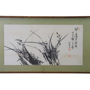 May include: A framed black and white ink painting of tall grass with a few small flowers. The painting is on a white background and has a green mat. The frame is brown wood. The painting has Chinese characters in black ink on the right side.