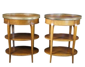 May include: Two oval-shaped wooden end tables with brass trim on the top shelf. Each table has three shelves and stands on four legs.