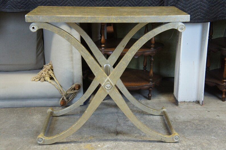K&ouml;nnte beinhalten: Ein goldfarbener Metalltisch mit rechteckiger Tischplatte und einem X-f&ouml;rmigen Gestell. Der Tisch hat eine Used-Look-Oberfl&auml;che und ist aus Metall.